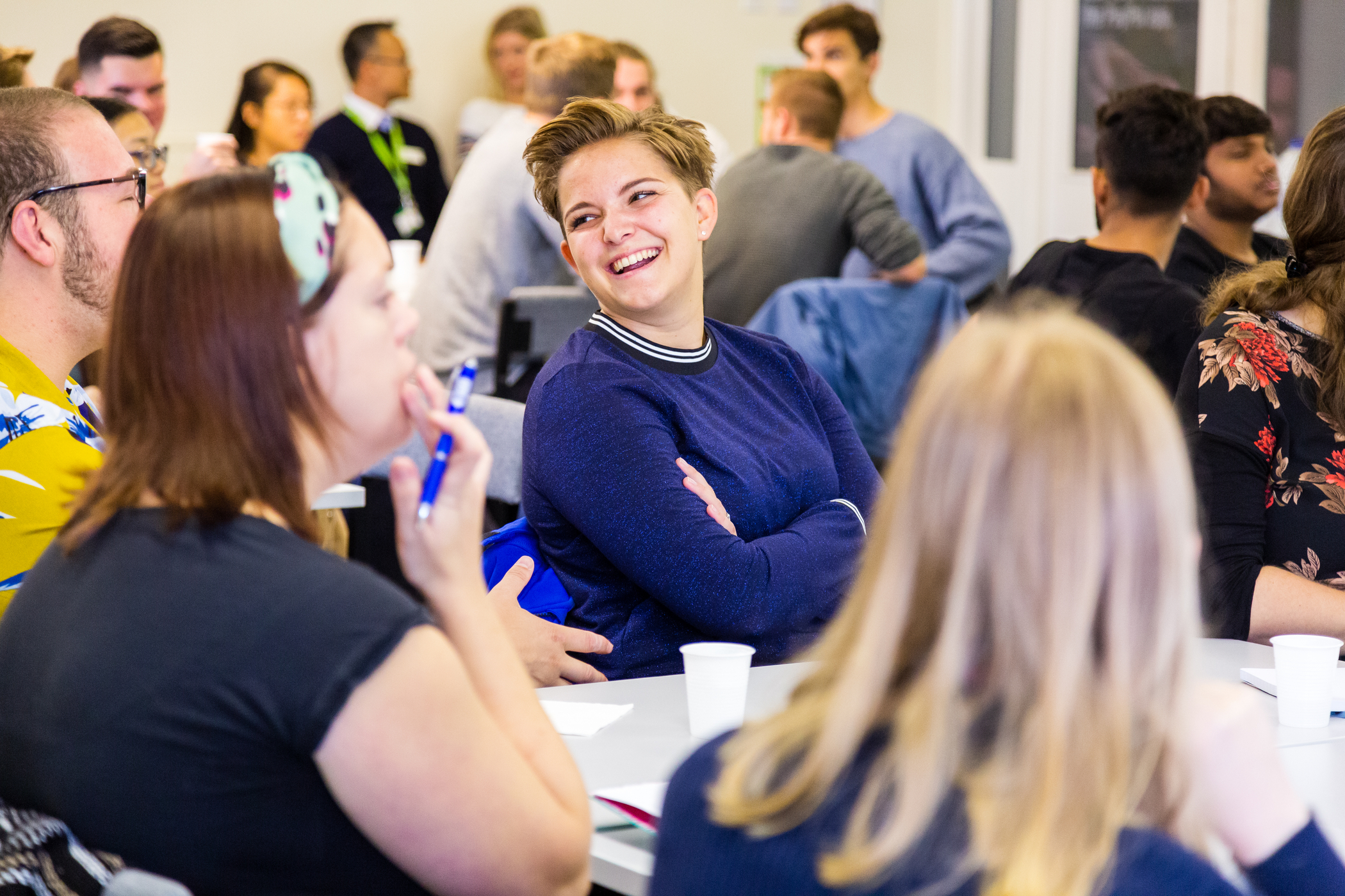 A woman sits in a crowded room smiling at a classmate.